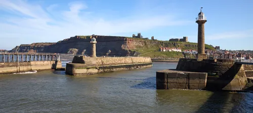 Whitby Abbey St Marys church and harbour from piers This landscape photograph shows Whitby Harbour in Yorkshire during a clear spring afternoon. The scene prominently features the two lighthouses at the entrance of the harbour, with the historic ruins of Whitby Abbey and St Mary’s Church visible on the hilltop in the background. The sunlight highlights the stone structure of the piers and lighthouses, while the houses of Whitby are clustered along the shoreline. St Mary’s Church stands out amongst the buildings on the green hillside, and Whitby Abbey’s ruins provide a dramatic focal point above the town. The image captures the maritime character of Whitby, showcasing the coastal heritage of Yorkshire with its harbour and lighthouse, and the distinctive architecture of the church and abbey overlooking the waterfront.